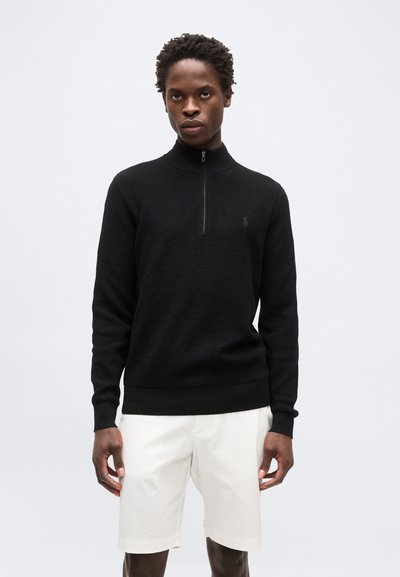 Homme portant un pull noir texturé à manches longues avec une fermeture éclair quart de longueur et un short blanc, debout devant un fond clair uni.
