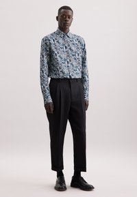 Camicia a maniche lunghe con motivo floreale in blu e bianco, abbinata a pantaloni neri a vita alta. Modello che indossa scarpe nere, in piedi su uno sfondo neutro.