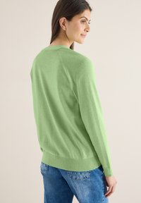 Femme tournée vers la droite portant un pull vert clair et un jean bleu, regardant légèrement par-dessus son épaule sur un fond clair uni.