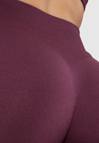 Vysoce pasové legíny v hluboké burgundské barvě, vyrobené z hladké, pružné tkaniny se bezešvým designem a mírným leskem, který zvýrazňuje kontury.