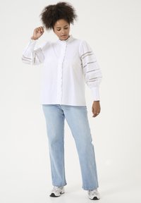 Blouse en coton blanc avec des détails en volants, un col montant et des manches bouffantes avec des inserts en dentelle. Portée avec un jean bleu clair et des baskets blanches.
