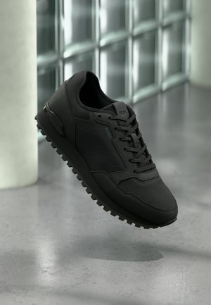 Schwarzer BOSS Sneaker mit strukturiertem Stoff, Schnürsenkeln und robuster Sohle, schwebend in der Luft vor grauem Boden und Wand aus Glasbausteinen.
