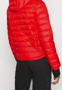 Chaqueta acolchada roja con capucha, diseño acolchado, textura brillante, puños elásticos y dobladillo que llega a la cintura. Combinada con pantalones negros.