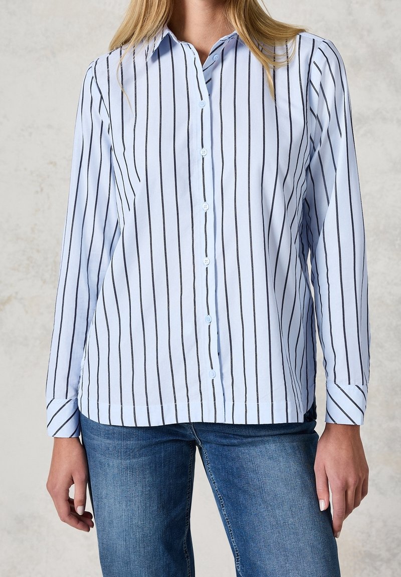 Femme portant une chemise bleu clair à manches longues avec des rayures verticales noires et un jean bleu, debout devant un fond neutre.