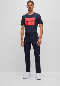 Navy short-sleeve t-shirt with a red rectangular graphic featuring "HUGO" in black, paired with navy trousers and white sneakers.