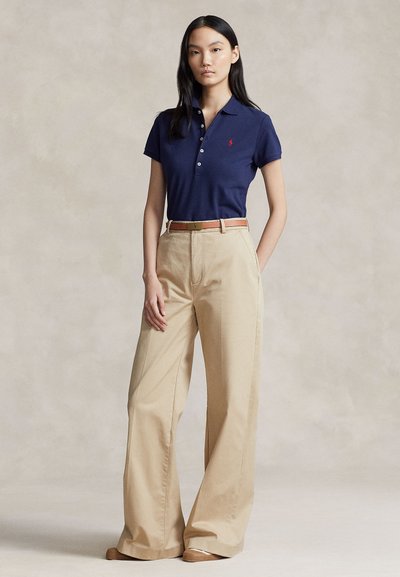 Ung kvinne stående iført marineblå poloskjorte, beige vidbukser med høy midje og brunt belte på nøytral bakgrunn.