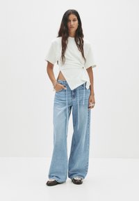 Weißes Wickeloberteil mit kurzen Ärmeln, kombiniert mit hellblauen weit geschnittenen Jeans. Lässiges Outfit mit bequemem Sitz und minimalen Details.