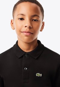 Schwarzes Polohemd mit strukturierter Oberfläche, ausgestattet mit einer Knopfleiste mit zwei Knöpfen und einem kleinen grünen gestickten Krokodil-Logo.