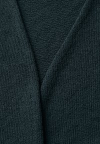 Tummanvihreä teksturoitu kangas pehmeällä, uritetulla pinnalla, jossa on V-mallinen kaula-aukko, tarjoten kerroksellisen ilmeen.