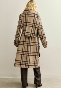 Blond person som bär en beige och brun rutigt lång kappa med matchande scarf, mörkbruna byxor och svarta klackskor, vänd mot väggen.