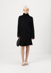 Black, textured sweater with a high neck and long sleeves paired with a matching black skirt. Holds a black bag, wearing beige sandals.