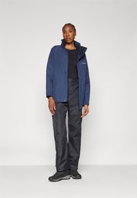 Navy weather-resistant jacket with a high collar and front buttons, paired with black waterproof pants and sturdy black boots.