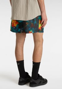 Farbige Batik-Shorts in Schwarz, mit leuchtenden Mustern in Orange, Grün und Blau; getragen mit schwarzen Socken und schwarzen Schuhen.