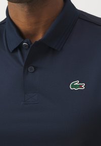 Mężczyzna noszący granatową koszulkę polo z dwoma guzikami i zielonym logo krokodyla na piersi.