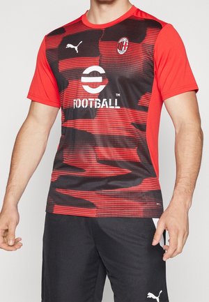 Mężczyzna w czerwono-czarnej koszulce piłkarskiej AC Milan z logo Puma i czarnych szortach Puma.