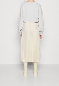 Sudadera gris claro de gran tamaño, falda midi de crema con pliegues y botas blancas hasta la rodilla, todo contra un fondo neutro.