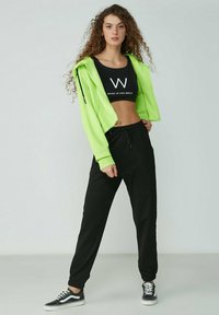 Neon grüne Cropped-Hoodie, schwarzes Cropped-Top mit Druck, schwarze Jogginghose und schwarze Sneaker. Weicher Stoff mit lockerer Passform, elastische Bündchen.