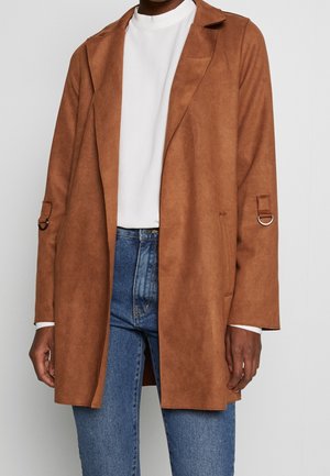 Osoba ubrana w brązowy zamszowy płaszcz z otwartym przodem, na białym topie z wysokim kołnierzem i niebieskich dżinsach na jednolitym białym tle.