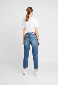 Mujer con coleta, vistiendo una camiseta blanca, jeans azules con un parche de logo en la parte trasera de la cinturilla, y zapatillas altas blancas, de pie frente a un fondo liso.