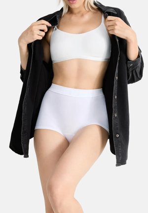 Vrouw draagt witte high-waist onderbroek en een witte beha, houdt een zwart denimjack open tegen een effen achtergrond.