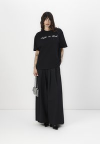 Črn oversized t-shirt z vezenjem "Café de Köl", v kombinaciji s širokima črnima hlačami in majhno torbico s srebrnimi bleščečimi detalji.