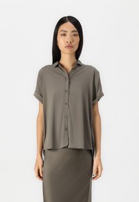 Camicia a maniche corte con colletto di color verde oliva, realizzata in tessuto liscio, con bottoni frontali e maniche arrotolate, abbinata a una gonna coordinata.