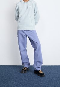 Ljusblå fleecehoodie med dragkedja och ficka, i kombination med lavendelfärgade bomullsbukser. Skorna har en svart sula med ett distinkt mönster.