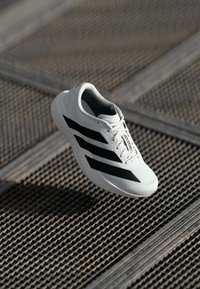 White running shoe with black stripes and "ADIZERO" text, floating above a textured metal grate surface.