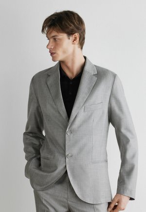 Junger Mann in einem hellgrauen Anzug und schwarzem Hemd, steht mit einer Hand in der Tasche und schaut nach links vor einem schlichten weißen Hintergrund.