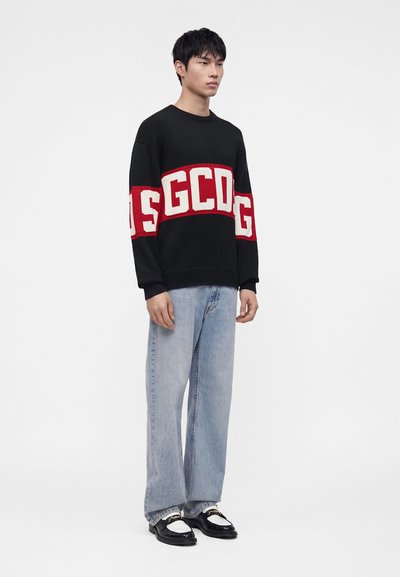 Jeune homme portant un pull noir avec des lettres rouges et blanches, un jean bleu clair et des chaussures noires et blanches, debout sur un fond uni.