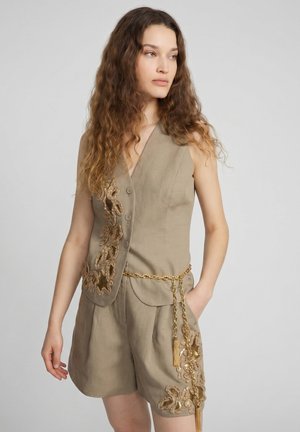 Femme aux longs cheveux ondulés portant un gilet beige sans manches et un short assorti avec une broderie florale dorée et une ceinture chaîne dorée.