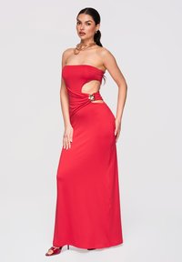 Femme debout portant une robe rouge sans bretelles avec découpe latérale et accent doré, associée à des talons rouges et un collier doré sur un fond uni.