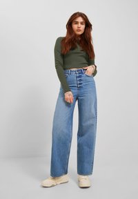 Zeleni uski longsleeve top spojen s visokim strukom i širokim plavim trapericama. Teksturirani svijetloplavi traper i kremasti tenisice upotpunjuju izgled.