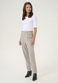 Haut ajusté blanc, pantalon beige clair sur mesure avec des plis et botas noires à la cheville. La tenue présente une texture lisse et un design minimaliste.