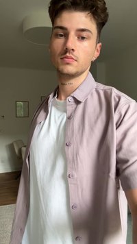 Lichtpaarse overhemd met knopen over een witte T-shirt, met een relaxte pasvorm en een zachte textuur. Achtergrond toont een neutrale kamer met meubels.
