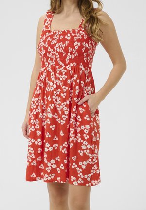 Rode bloemenjurk met geplooide taille, vierkante halslijn en brede bandjes. Voorzien van witte bloemmotieven en zijzakken.