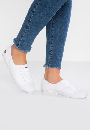 Pies con zapatillas blancas y jeans ajustados azul con dobladillo deshilachado sobre un fondo blanco liso.