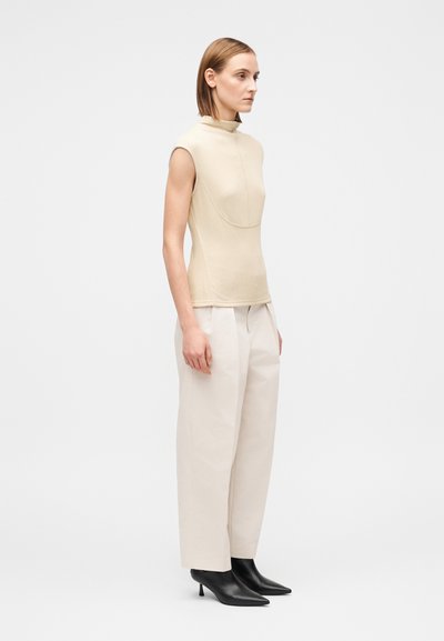 Beige ärmlös topp med hög krage, kombinerad med krämfärgade byxor med hög midja och svarta spetsiga klackskor. Slät textur, skräddarsydd passform.