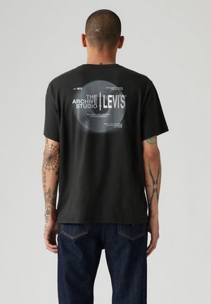 Hombre con los brazos tatuados que lleva una camiseta negra de Levi's con un gráfico circular blanco y el texto "The Archive Studio" en la parte trasera, combinada con vaqueros azules.
