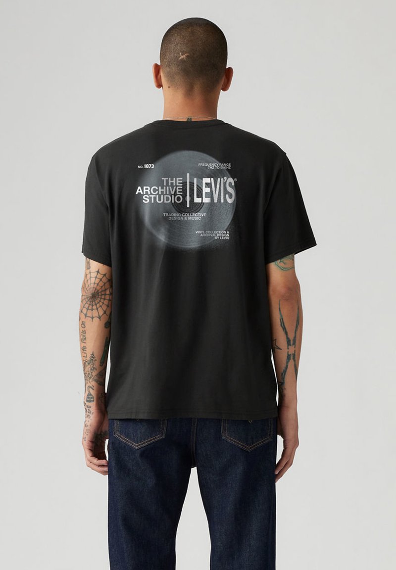 Homme aux bras tatoués portant un T-shirt noir Levi's avec un graphisme circulaire blanc et le texte "The Archive Studio" au dos, associé à un jean bleu.