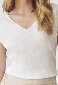 Biały t-shirt z dekoltem w serek, z krótkim rękawem oraz fakturowanym, lekko przejrzystym materiałem. Posiada delikatne przeszycia wzdłuż dekoltu.