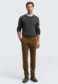 Maglione grigio scuro con scollatura a girocollo, abbinato a pantaloni marroni e scarpe nere. L'outfit è casual e ben aderente.