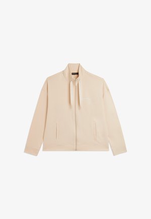 Veste beige zippée avec col haut, deux poches avant, cordons de marque et logo discret sur la poitrine.
