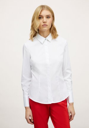 Camisa blanca de manga larga con botones, de cuello puntiagudo y textura suave, presentada junto a pantalones rojos.