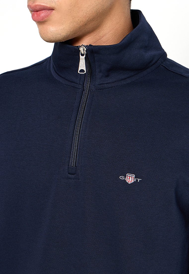 Dunkelblaues Fleece-Half-Zip-Pullover mit einem silbernen Reißverschluss, hohem Kragen und kleinem gesticktem Logo auf der linken Brust. Weiche Textur.