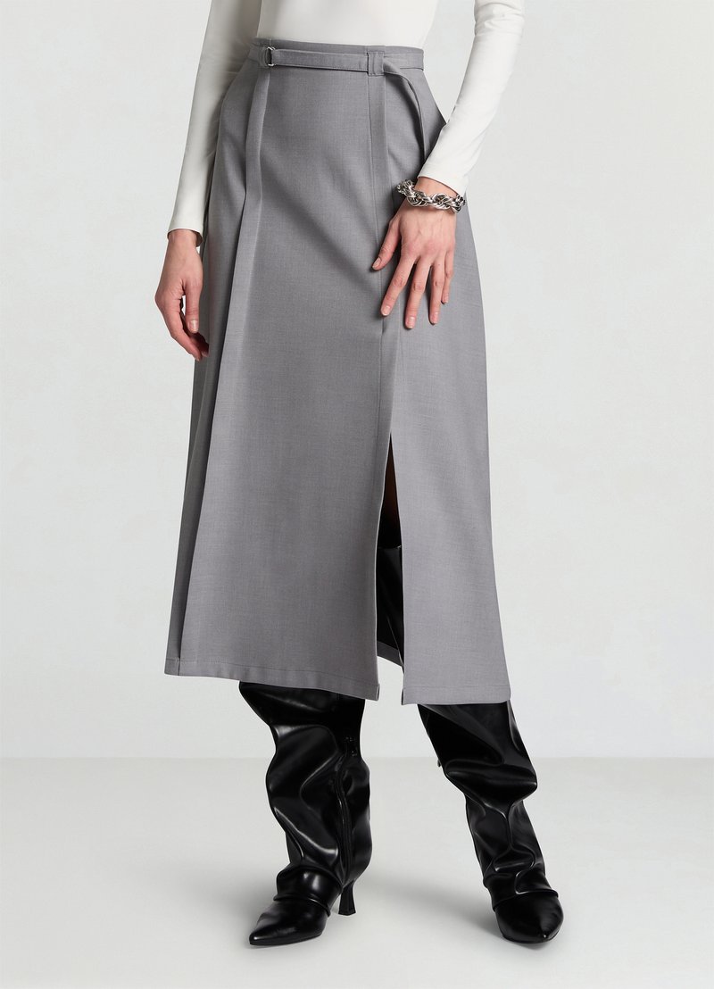 Jupe midi grise avec un détail ceinture, ornée de plis et d'une fente latérale. Associée à des cuissardes noires brillantes. Texture de tissu lisse et formel.