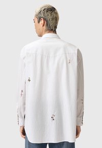 Λευκό πουκάμισο με ρίγες, διακοσμημένο με κεντημένα λουλουδένια σχέδια στην πλάτη και τα μανίκια, με κλασικό γιακά και κουμπώματα.