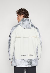 White outdoor jacket featuring a gray marble design on the shoulders and hood, with a logo at the hem. Includes a large back pocket.