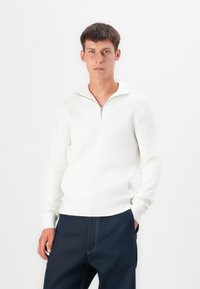 Suéter blanco de canalé con cuello de media cremallera y mangas largas, combinado con pantalones azul oscuro. La tela parece suave y con textura.