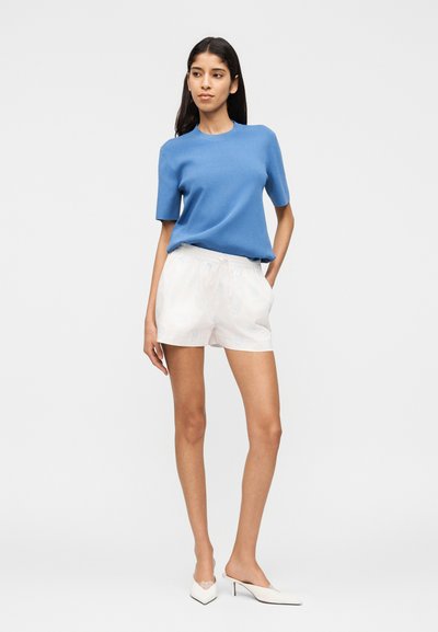 Γυναίκα που ποζάρει με μπλε κοντομάνικο τοπ, λευκά σορτς με λουλουδάτο μοτίβο και λευκά μυτερά τακούνια, σε απλό φωτεινό φόντο.